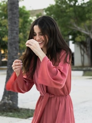 A woman with long dark hair is wearing a flowing, pink robe-style dress. She appears to be outdoors in a natural setting with trees in the background. Her expression is joyful, and she is gesturing with her hands in motion.