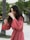 A woman with long dark hair is wearing a flowing, pink robe-style dress. She appears to be outdoors in a natural setting with trees in the background. Her expression is joyful, and she is gesturing with her hands in motion.
