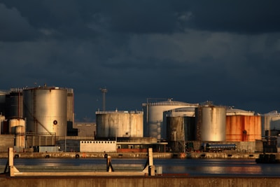 A series of large industrial storage tanks made of metal are lined up along a waterfront. The tanks have varied rust and wear, with shadows cast by the low sun under dark, overcast skies. The scene has a stark industrial appearance.