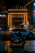 A nighttime urban scene featuring a vibrant, neon-lit gate with glowing orange lights. Several flags are displayed on top of the gate. A car passes beneath the gate, while in the foreground, a person wearing a mask rides a scooter. The scene is bustling with activity, capturing a sense of movement across the street.