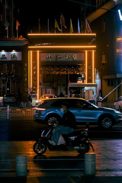 A nighttime urban scene featuring a vibrant, neon-lit gate with glowing orange lights. Several flags are displayed on top of the gate. A car passes beneath the gate, while in the foreground, a person wearing a mask rides a scooter. The scene is bustling with activity, capturing a sense of movement across the street.