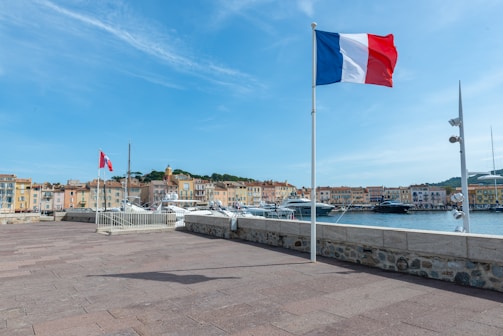 A waterfront scene featuring a French flag waving on a pole by the edge of a paved area. In the background, there are several boats docked at a marina and a row of colorful buildings. The sky is clear and blue, suggesting a sunny day.
