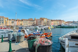 A picturesque harbor scene featuring colorful fishing boats docked alongside a vibrant waterfront. The foreground displays fishing nets and equipment, while the background shows a line of charming, pastel-colored buildings lining the harbor. The setting suggests a quaint coastal village on a bright, sunny day with a clear blue sky.
