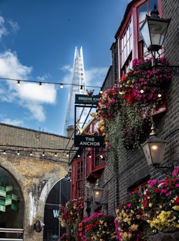 A vibrant street scene showing a traditional pub with a sign reading 'The Anchor.' The building is adorned with colorful flower baskets and classic-style lanterns. The background features a brick railway arch and a modern skyscraper with a pointed top, set against a partially cloudy blue sky.