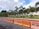 A row of orange traffic barriers lines the side of a road adjacent to a fence. Behind the fence, well-maintained landscaping features palm trees and various shrubs. The sky above is partly cloudy, with patches of blue visible.