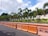 A row of orange traffic barriers lines the side of a road adjacent to a fence. Behind the fence, well-maintained landscaping features palm trees and various shrubs. The sky above is partly cloudy, with patches of blue visible.