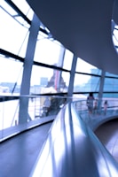 A modern architectural interior featuring a curved metal railing in the foreground, with expansive glass windows creating a bright and airy atmosphere. Blurred figures in the background suggest people walking along a curved pathway.