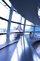 A modern architectural interior featuring a curved metal railing in the foreground, with expansive glass windows creating a bright and airy atmosphere. Blurred figures in the background suggest people walking along a curved pathway.