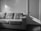 A gray upholstered sofa with three cushions stands against a wall. Sunlight casts geometric patterns on the wall and part of the sofa. A closed door with a handle is visible on the right side of the image. The floor appears to be dark wood.