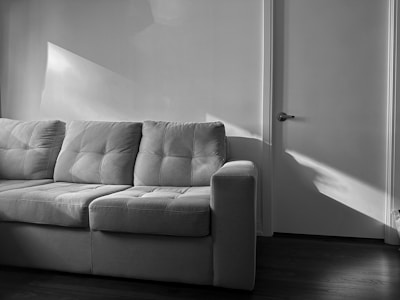 A gray upholstered sofa with three cushions stands against a wall. Sunlight casts geometric patterns on the wall and part of the sofa. A closed door with a handle is visible on the right side of the image. The floor appears to be dark wood.