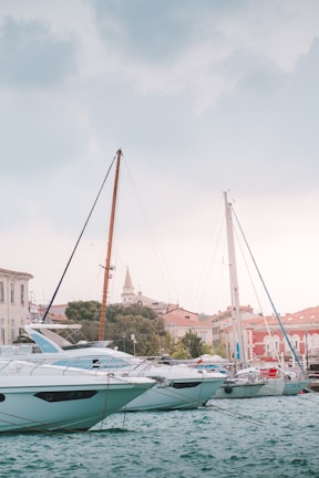 Several yachts are docked in a marina with calm turquoise waters. The background features an array of buildings with red-tiled roofs, and a church tower is prominently visible. The sky is overcast with soft hints of blue and gray clouds.