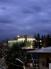 A large building is illuminated in the evening with neon green and yellow lights. It is surrounded by trees and appears to be a hotel or inn as indicated by the signage on the rooftop. The sky is overcast and dark, creating a moody, nocturnal atmosphere. In the foreground, palm trees and a rooftop with garments hanging can be seen.