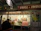 A food stall named 'Grill Imbiss' offers a variety of grilled sausages and hot dogs, with a menu displaying images and prices. The interior is decorated with bottles and packages arranged neatly. A man is standing in front of the counter, and a red and white checkered cloth covers part of the counter. There are promotional posters and price lists for drinks like Glühwein and coffee.