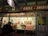A food stall named 'Grill Imbiss' offers a variety of grilled sausages and hot dogs, with a menu displaying images and prices. The interior is decorated with bottles and packages arranged neatly. A man is standing in front of the counter, and a red and white checkered cloth covers part of the counter. There are promotional posters and price lists for drinks like Glühwein and coffee.