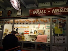 A food stall named 'Grill Imbiss' offers a variety of grilled sausages and hot dogs, with a menu displaying images and prices. The interior is decorated with bottles and packages arranged neatly. A man is standing in front of the counter, and a red and white checkered cloth covers part of the counter. There are promotional posters and price lists for drinks like Glühwein and coffee.