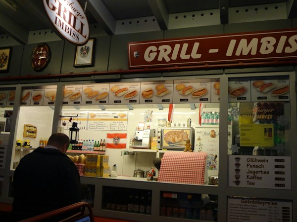 A food stall named 'Grill Imbiss' offers a variety of grilled sausages and hot dogs, with a menu displaying images and prices. The interior is decorated with bottles and packages arranged neatly. A man is standing in front of the counter, and a red and white checkered cloth covers part of the counter. There are promotional posters and price lists for drinks like Gl&uuml;hwein and coffee.