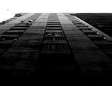 A black and white photograph of a tall residential building taken from a low angle, emphasizing its height and structure. The stark contrast highlights the windows and balconies. A lone bird is visible flying in the overcast sky, adding a sense of scale and dynamism to the otherwise static scene.