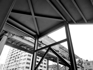 A monochrome architectural image featuring intersecting steel beams and structures, with a high-rise building in the background. The composition includes a pedestrian bridge partially visible with intricate shadows and lines formed by the framework.