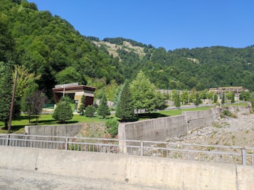 A lush green landscape with dense forested hills and a clear blue sky. In the foreground, there is a grassy area with several trees and a modern building, possibly a hotel. Concrete barriers and a dry riverbed are also visible.