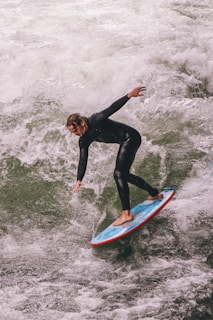 A person in a black wetsuit surfing on a wave, expertly balancing on a blue surfboard with a red trim. The white foam of the churning water contrasts against the vibrant colors of the surfboard.
