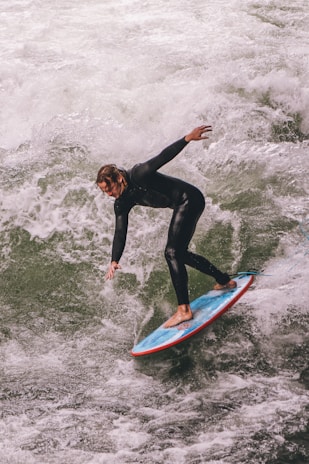 A person in a black wetsuit surfing on a wave, expertly balancing on a blue surfboard with a red trim. The white foam of the churning water contrasts against the vibrant colors of the surfboard.