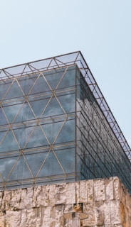 A modern architectural structure features a geometric metal lattice over a glass facade with a stone foundation. The lattice creates a grid pattern, adding an industrial and contemporary aesthetic. The stone base has a rough texture, contrasting with the sleek upper levels. A cylindrical outdoor lamp is visible near the bottom.
