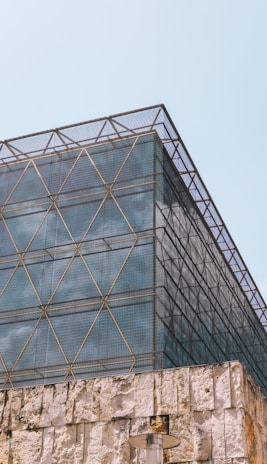 A modern architectural structure features a geometric metal lattice over a glass facade with a stone foundation. The lattice creates a grid pattern, adding an industrial and contemporary aesthetic. The stone base has a rough texture, contrasting with the sleek upper levels. A cylindrical outdoor lamp is visible near the bottom.