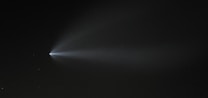 A bright object with a long, luminous tail seems to be moving through a dark night sky. The object resembles a comet or rocket, emitting a trail of light that gradually fades into the darkness. Surrounding the glowing trail are faint stars scattered across the background.