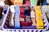A basket containing neatly arranged stacks of colorful ceramic tiles in various shades such as blue, red, yellow, and white, with some featuring intricate patterns. The basket itself has a purple and white woven design.