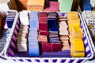 A basket containing neatly arranged stacks of colorful ceramic tiles in various shades such as blue, red, yellow, and white, with some featuring intricate patterns. The basket itself has a purple and white woven design.
