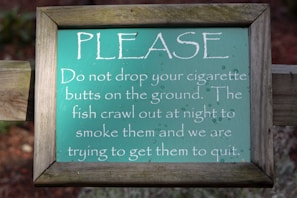 A wooden-framed sign with a humorous message about not dropping cigarette butts. The sign is green with white text, featuring a playful comment about fish smoking the butts and an attempt to get them to quit.