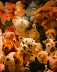 A variety of plush stuffed animals, including dogs, horses, and bears, are piled together in different colors such as brown, white, and pink. Some of these toys have tags attached to them, indicating they are from a store or collection.