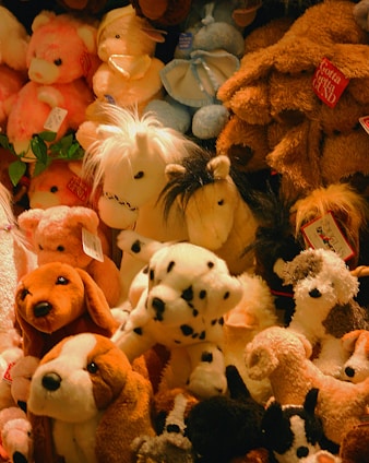 A variety of plush stuffed animals, including dogs, horses, and bears, are piled together in different colors such as brown, white, and pink. Some of these toys have tags attached to them, indicating they are from a store or collection.