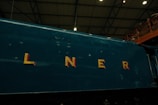 The side of a dark green train engine is shown with large, bold yellow letters spelling 'LNER' emblazoned across it. The background features a high ceiling with a metal truss structure, and part of an observation deck with railings, where a few people are visible.