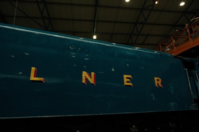 The side of a dark green train engine is shown with large, bold yellow letters spelling 'LNER' emblazoned across it. The background features a high ceiling with a metal truss structure, and part of an observation deck with railings, where a few people are visible.