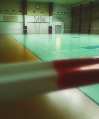 A spacious indoor gymnasium with a green and brown floor designed for sports like basketball or volleyball. The room is empty and has minimal ambient lighting, with large beams and walls painted in light colors. A basketball hoop is mounted on the wall, and there is a clock visible above it. Red and white barrier tape is stretched across the room, likely indicating a restricted area.