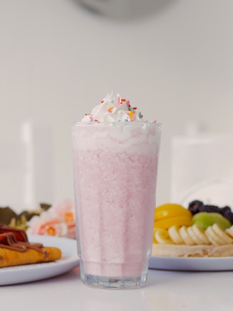 A tall glass filled with a pink milkshake, topped with whipped cream and colorful sprinkles, stands prominently in the foreground. In the background, there is a plate with a variety of fresh fruits including bananas, grapes, and sliced peaches, and another plate with a dessert topped with chocolate.