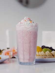 A tall glass filled with a pink milkshake, topped with whipped cream and colorful sprinkles. In the background, plates of assorted fruits, including bananas, grapes, and sliced fruits, are visible along with some pink flowers.