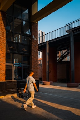 A young person walks through an urban street setting with modern architecture. The scene features brick walls and glass windows reflecting the warm glow of the setting sun. Shadows are cast on the pavement, creating dynamic lines.