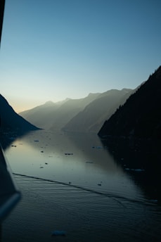 A serene water body is flanked by misty, rugged mountains under a clear sky. Small chunks of ice float on the water's surface, which reflects the sky and mountains. The scene conveys a sense of calm and solitude.