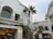 An outdoor shopping area featuring white stucco buildings with arched windows and decorative iron railings. A tall palm tree stands prominently in the center, surrounded by greenery and string lights. The area appears lively, with people visible in the background.