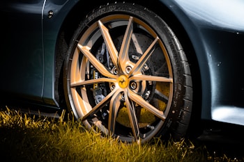 A close-up view of an elegant and sporty alloy wheel of a luxury car, with a detailed focus on its sleek, multi-spoke design and the manufacturer's emblem at the center. The wheel is surrounded by a high-performance tire, and the car's glossy exterior is visible. Sunlight casts vibrant reflections, highlighting the metallic finish, while the foreground features lush green grass.