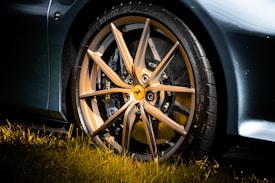 A close-up view of an elegant and sporty alloy wheel of a luxury car, with a detailed focus on its sleek, multi-spoke design and the manufacturer's emblem at the center. The wheel is surrounded by a high-performance tire, and the car's glossy exterior is visible. Sunlight casts vibrant reflections, highlighting the metallic finish, while the foreground features lush green grass.