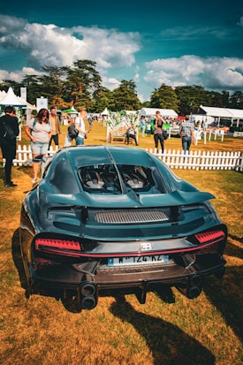 A luxury sports car is parked on a grassy area surrounded by people. The vehicle is sleek and modern with a shiny black exterior. In the background, a small crowd is gathered, with people standing near white picket fences and tents. The scene is set outdoors with trees and a partly cloudy sky.