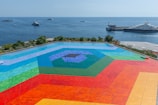 A vibrant, multicolored geometric design covers a large, open area on the ground. The design features concentric hexagons in shades of blue, green, red, orange, and yellow. In the background, there is a clear blue ocean with several yachts and a docked ship, framed by lush green vegetation.