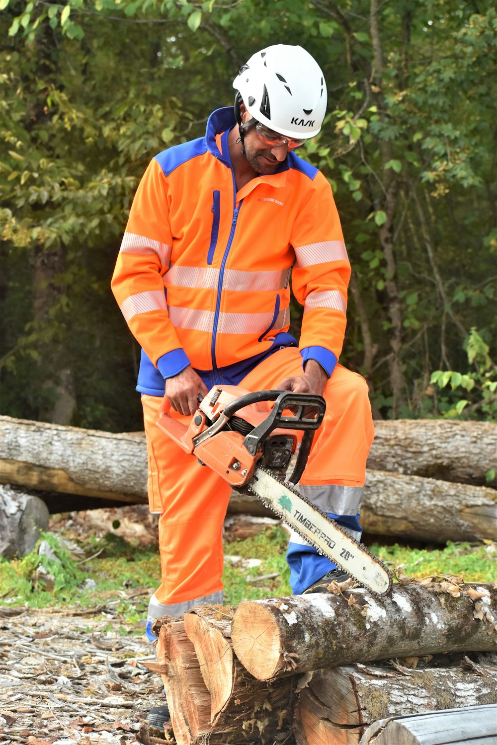 Un hombre con una chaqueta naranja y azul está cortando un tronco