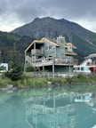 A wooden cabin with a rustic design is situated by a calm body of water, reflecting the building's structure on the surface. The cabin is surrounded by lush greenery, with a dense forest and towering mountain in the background. The sky is overcast, adding a serene and tranquil feel to the scene.