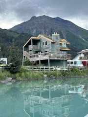 A wooden cabin with a rustic design is situated by a calm body of water, reflecting the building's structure on the surface. The cabin is surrounded by lush greenery, with a dense forest and towering mountain in the background. The sky is overcast, adding a serene and tranquil feel to the scene.