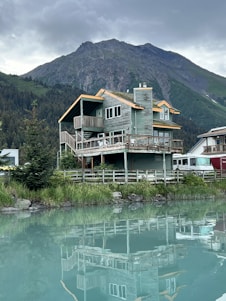 A wooden cabin with a rustic design is situated by a calm body of water, reflecting the building's structure on the surface. The cabin is surrounded by lush greenery, with a dense forest and towering mountain in the background. The sky is overcast, adding a serene and tranquil feel to the scene.