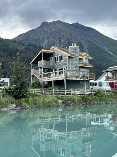 A wooden cabin with a rustic design is situated by a calm body of water, reflecting the building's structure on the surface. The cabin is surrounded by lush greenery, with a dense forest and towering mountain in the background. The sky is overcast, adding a serene and tranquil feel to the scene.